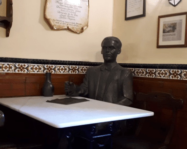 Entre azulejos decorativos y cuadros enmarcados, una estatua de bronce reposa elegantemente en una mesa de café. Ideal para reflexionar durante "Federico García Lorca: Tour guiado por Granada", que captura la esencia artística de Lorca. - ForTwo - Planes para parejas