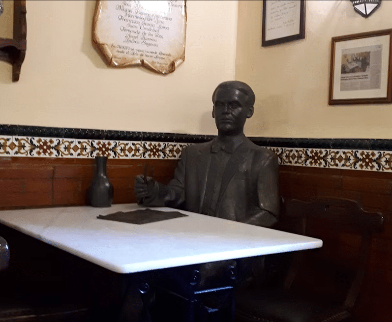 Entre azulejos decorativos y cuadros enmarcados, una estatua de bronce reposa elegantemente en una mesa de café. Ideal para reflexionar durante "Federico García Lorca: Tour guiado por Granada", que captura la esencia artística de Lorca. - ForTwo - Planes para parejas