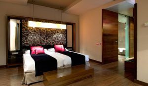 El Hotel Vincci Soho de Madrid ofrece una moderna habitación con dos camas, detalles en rojo y negro, cabecero floral, suelo de madera y baño con puerta abierta. Ubicado en el vibrante corazón de la ciudad, combina un diseño elegante con un ambiente cálido para una experiencia acogedora. - ForTwo - Planes para parejas