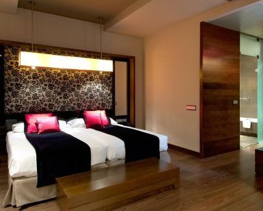 El Hotel Vincci Soho de Madrid ofrece una moderna habitación con dos camas, detalles en rojo y negro, cabecero floral, suelo de madera y baño con puerta abierta. Ubicado en el vibrante corazón de la ciudad, combina un diseño elegante con un ambiente cálido para una experiencia acogedora. - ForTwo - Planes para parejas