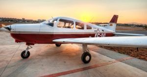 Una pequeña avióneta roja y blanca está estacionada en una pista al atardecer, lista para un Vuelo de iniciación en avióneta por Montserrat (Barcelona) sobre el impresionante paisaje. - ForTwo - Planes para parejas