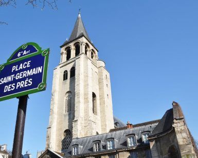 Placa de calle de la Place Saint-Germain des Prés con la torre de la iglesia al fondo bajo un cielo azul claro, que recuerda el encanto que se encuentra en un "Recorrido guiado por el Barrio Latino" gratuito en París. - ForTwo - Planes para parejas
