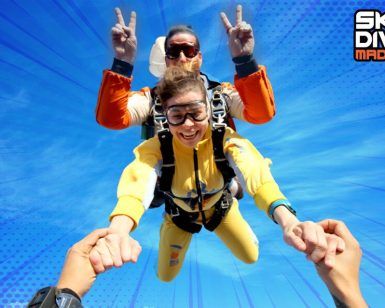 Paracaidistas tándem con vibrantes trajes amarillos se elevan alegremente con su instructor bajo un cielo azul despejado. ¡Vive la emoción del "Paracaidismo tándem desde 4000 metros" en SkyDive Madrid!. - ForTwo - Planes para parejas