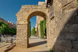 Arco de piedra y muralla antigua en una zona histórica de la ciudad, que recuerdan las vistas del "Free tour por la Barcelona romana", con árboles y gente al fondo. - ForTwo - Planes para parejas