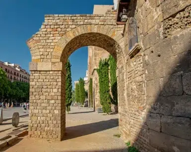 Arco de piedra y muralla antigua en una zona histórica de la ciudad, que recuerdan las vistas del "Free tour por la Barcelona romana", con árboles y gente al fondo. - ForTwo - Planes para parejas