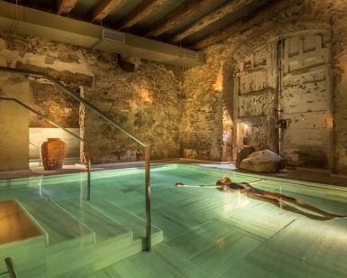 En una piscina de hidromasaje cubierta con poca luz, paredes de piedra y decoración rústica, uno puede disfrutar del relajante lujo del "Baño termal con aromaterapia y masaje de 15 minutos por Aire de Vallromanes" en Barcelona. - ForTwo - Planes para parejas