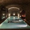 Dos personas flotan serenamente en la piscina cubierta tenuemente iluminada de Aires de Barcelona: Baño termal, rodeada de paredes de ladrillo y un techo elegantemente arqueado, que captura la esencia tranquila de una experiencia de baño termal tradicional. - ForTwo - Planes para parejas
