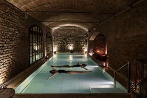 Dos personas flotan serenamente en la piscina cubierta tenuemente iluminada de Aires de Barcelona: Baño termal, rodeada de paredes de ladrillo y un techo elegantemente arqueado, que captura la esencia tranquila de una experiencia de baño termal tradicional. - ForTwo - Planes para parejas