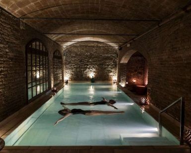Dos personas flotan serenamente en la piscina cubierta tenuemente iluminada de Aires de Barcelona: Baño termal, rodeada de paredes de ladrillo y un techo elegantemente arqueado, que captura la esencia tranquila de una experiencia de baño termal tradicional. - ForTwo - Planes para parejas