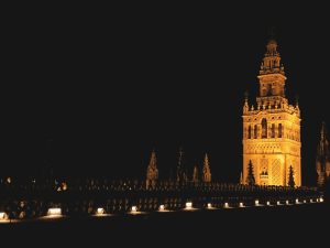 La torre gótica iluminada se eleva contra el oscuro cielo nocturno, sus detalles ornamentados bañados de luz, recuerdan el resplandor sereno de un Hammam sencillo en Aire de Sevilla. - ForTwo - Planes para parejas