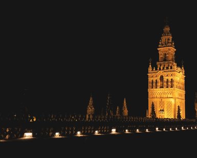 La torre gótica iluminada se eleva contra el oscuro cielo nocturno, sus detalles ornamentados bañados de luz, recuerdan el resplandor sereno de un Hammam sencillo en Aire de Sevilla. - ForTwo - Planes para parejas