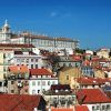 Bajo el vibrante cielo azul, edificios con tejados rojos rodean una histórica iglesia blanca con su cúpula, capturando la esencia de Alfama, Lisboa. Únete al "Tour gratuito por Alfama (Lisboa)" para explorar este pintoresco barrio lleno de encanto e historia. - ForTwo - Planes para parejas