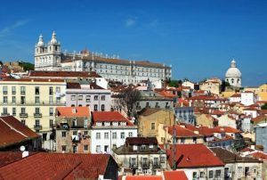 Bajo el vibrante cielo azul, edificios con tejados rojos rodean una histórica iglesia blanca con su cúpula, capturando la esencia de Alfama, Lisboa. Únete al "Tour gratuito por Alfama (Lisboa)" para explorar este pintoresco barrio lleno de encanto e historia. - ForTwo - Planes para parejas