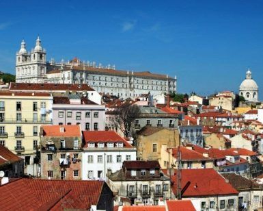 Bajo el vibrante cielo azul, edificios con tejados rojos rodean una histórica iglesia blanca con su cúpula, capturando la esencia de Alfama, Lisboa. Únete al "Tour gratuito por Alfama (Lisboa)" para explorar este pintoresco barrio lleno de encanto e historia. - ForTwo - Planes para parejas