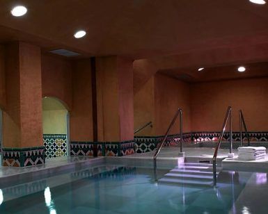 Como parte de la "OFERTA: Circuito en Baños Árabes para 2 con cava, bombones y masaje opcional", la piscina cubierta con poca luz cuenta con escalones rodeados de cálidas paredes marrones y azulejos decorativos. - ForTwo - Planes para parejas
