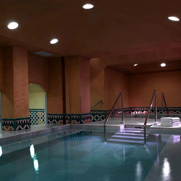Como parte de la "OFERTA: Circuito en Baños Árabes para 2 con cava, bombones y masaje opcional", la piscina cubierta con poca luz cuenta con escalones rodeados de cálidas paredes marrones y azulejos decorativos. - ForTwo - Planes para parejas