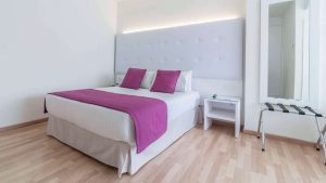 Disfrute de una habitación moderna con cama doble, ropa de cama blanca y morada, cabecero alto y mesita de noche en Escapada en la Costa Blanca del hotel Albahia (Alicante). Disfrute del encanto de Alicante cerca de las resplandecientes costas de la Costa Blanca. - ForTwo - Planes para parejas