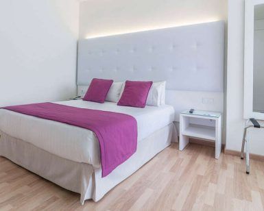 Disfrute de una habitación moderna con cama doble, ropa de cama blanca y morada, cabecero alto y mesita de noche en Escapada en la Costa Blanca del hotel Albahia (Alicante). Disfrute del encanto de Alicante cerca de las resplandecientes costas de la Costa Blanca. - ForTwo - Planes para parejas