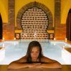 Una mujer se relaja junto a una piscina cubierta iluminada con los ojos cerrados, rodeada de azulejos decorativos y arcos, evocando el ambiente sereno del Baño sencillo en Aljibe Baños Árabes de San Miguel. - ForTwo - Planes para parejas