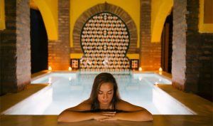 Una mujer se relaja junto a una piscina cubierta iluminada con los ojos cerrados, rodeada de azulejos decorativos y arcos, evocando el ambiente sereno del Baño sencillo en Aljibe Baños Árabes de San Miguel. - ForTwo - Planes para parejas