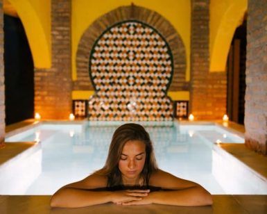 Una mujer se relaja junto a una piscina cubierta iluminada con los ojos cerrados, rodeada de azulejos decorativos y arcos, evocando el ambiente sereno del Baño sencillo en Aljibe Baños Árabes de San Miguel. - ForTwo - Planes para parejas