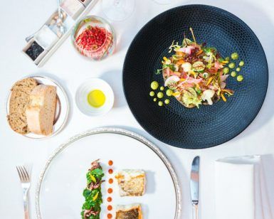 Un elegante montaje gastronómico para el "Masaje y almuerzo ejecutivo a la carta en el Hotel InterContinental Gran Vía (Madrid)" presenta dos platos gourmet, pan, aceite de oliva y condimentos sobre un mantel blanco impecable, perfecto para una experiencia refinada. - ForTwo - Planes para parejas