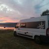 Una Catalunyavan, bajo un vibrante atardecer junto a un tranquilo lago con colinas a lo lejos, invita a explorar más allá de Barcelona. Ideal para quienes buscan "Alquiler de furgos camperizadas con Catalunyavan (Barcelona)" y sumergirse en la serena belleza de la naturaleza. - ForTwo - Planes para parejas