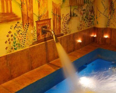 El agua fluye hacia una serena piscina azul, rodeada de un mural de edificios antiguos y naturaleza, creando una escapada tranquila que recuerda a la "Oferta Lunes a Viernes de sesión de Spa en Aqua Urban Spa Barcelona. - ForTwo - Planes para parejas