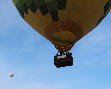 Captura un primer plano de un vibrante globo aerostático amarillo y verde en vuelo, con uno más pequeño detrás, contra el cielo azul claro. Disfruta de esta fascinante escena durante un vuelo en globo con Eolofly en Aranjuez sobre pintorescos paisajes. - ForTwo - Planes para parejas