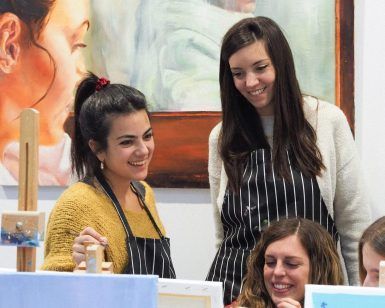 Tres mujeres con delantales de rayas disfrutan de un animado "Arte y Vino: Taller de Pintura y Vino" en Barcelona, sonriendo e interactuando entre sí. - ForTwo - Planes para parejas