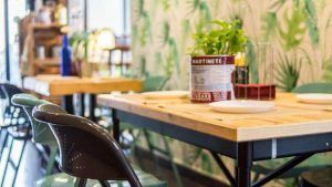 Esta moderna cafetería, con mesas de madera, sillas verdes y un centro de mesa con plantas, rezuma el ambiente del Restaurante Artefacto (Sevilla). Un papel pintado con estampado de hojas adorna las paredes, realzando el encantador ambiente de los cafés sevillanos. - ForTwo - Planes para parejas