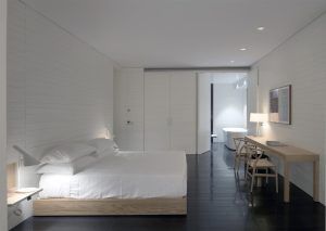 La habitación minimalista del Hotel Atrio (Cáceres) incluye cama, escritorio y silla. Tiene suelo oscuro y paredes blancas, con una serena vista al baño, combinando sencillez y elegancia. - ForTwo - Planes para parejas