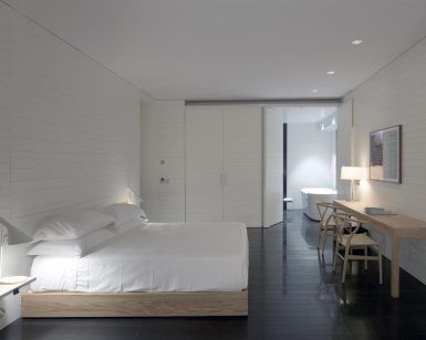 La habitación minimalista del Hotel Atrio (Cáceres) incluye cama, escritorio y silla. Tiene suelo oscuro y paredes blancas, con una serena vista al baño, combinando sencillez y elegancia. - ForTwo - Planes para parejas