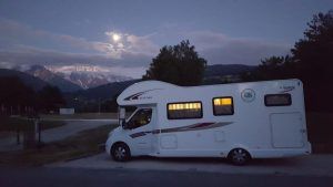 Una "Alquila una autocaravana en Barcelona" estacionada al anochecer, recortada contra majestuosas montañas y una luna llena, susurrando historias de aventuras. - ForTwo - Planes para parejas