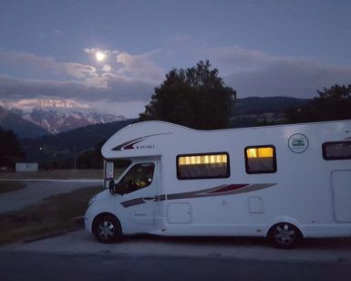 Una "Alquila una autocaravana en Barcelona" estacionada al anochecer, recortada contra majestuosas montañas y una luna llena, susurrando historias de aventuras. - ForTwo - Planes para parejas