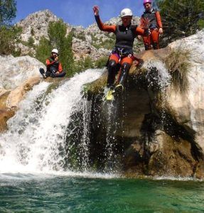 Una persona con casco salta desde una pequeña cascada a las cristalinas aguas de la Alpujarra, mientras otros dos observan, cautivados por esta emocionante experiencia de "Barranquismo en Granada-Poqueira, en la Alpujarra". - ForTwo - Planes para parejas