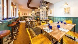 El restaurante Bache San Pedro (Sevilla) cuenta con acogedoras mesas de madera, cristalería azul vibrante, sillas amarillas y azulejos de pared decorativos que encarnan el encanto de Sevilla. - ForTwo - Planes para parejas