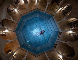 Una persona flota en una piscina octogonal en Baño en Pareja Hammam Al Ándalus Málaga Baños Árabes, rodeada de columnas e iluminación tenue, creando el ambiente sereno de los antiguos Baños Árabes. - ForTwo - Planes para parejas