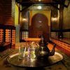 Un juego de té marroquí con una tetera de plata y vasos se encuentra en una bandeja en una habitación cálidamente iluminada con paredes estampadas, creando un ambiente de Baños Árabes en Granada similar a la experiencia del Tratamiento Sheik en Aljibe de San Miguel. - ForTwo - Planes para parejas