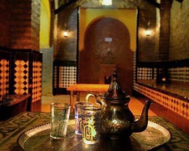 Un juego de té marroquí con una tetera de plata y vasos se encuentra en una bandeja en una habitación cálidamente iluminada con paredes estampadas, creando un ambiente de Baños Árabes en Granada similar a la experiencia del Tratamiento Sheik en Aljibe de San Miguel. - ForTwo - Planes para parejas