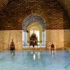 Una tranquila piscina de hidromasaje cubierta con arcos de ladrillo, paredes de mármol y faroles ornamentados proyecta sombras intrincadas, evocando la calma de una Sesión de baños árabes en Medina Mudéjar en Toledo. - ForTwo - Planes para parejas