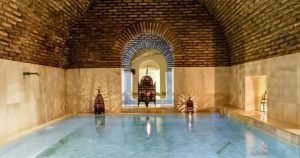 Una tranquila piscina de hidromasaje cubierta con arcos de ladrillo, paredes de mármol y faroles ornamentados proyecta sombras intrincadas, evocando la calma de una Sesión de baños árabes en Medina Mudéjar en Toledo. - ForTwo - Planes para parejas