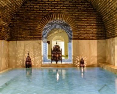 Una tranquila piscina de hidromasaje cubierta con arcos de ladrillo, paredes de mármol y faroles ornamentados proyecta sombras intrincadas, evocando la calma de una Sesión de baños árabes en Medina Mudéjar en Toledo. - ForTwo - Planes para parejas