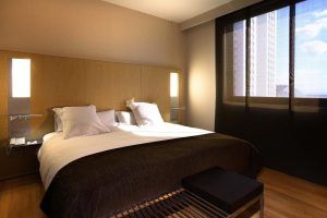 Experimente la comodidad en el Hotel Barceló (Valencia) con una moderna habitación que cuenta con una acogedora cama doble, un elegante cabecero de madera y un gran ventanal con elegantes cortinas negras. - ForTwo - Planes para parejas