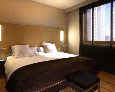 Experimente la comodidad en el Hotel Barceló (Valencia) con una moderna habitación que cuenta con una acogedora cama doble, un elegante cabecero de madera y un gran ventanal con elegantes cortinas negras. - ForTwo - Planes para parejas