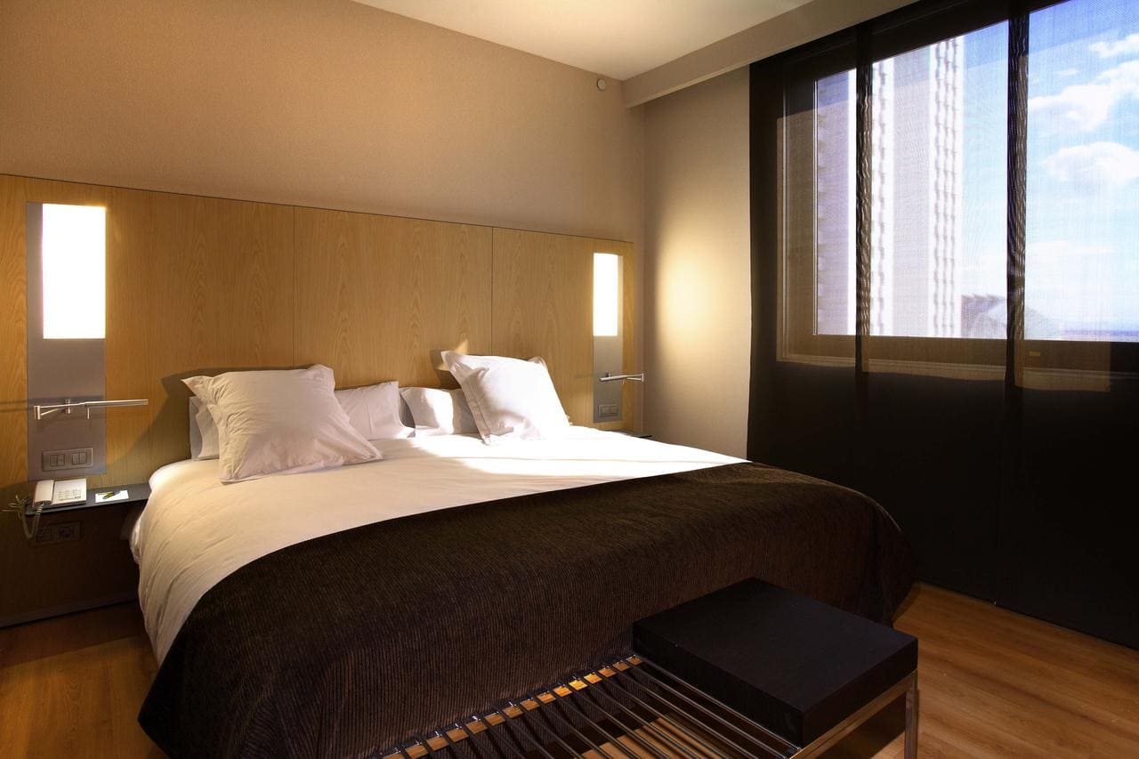 Experimente la comodidad en el Hotel Barceló (Valencia) con una moderna habitación que cuenta con una acogedora cama doble, un elegante cabecero de madera y un gran ventanal con elegantes cortinas negras. - ForTwo - Planes para parejas
