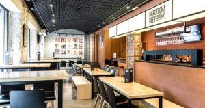El moderno interior de la cervecería cuenta con mesas y sillas de madera y una acogedora zona de bar. Los amplios ventanales iluminan el cartel de Barcelona Beer Company, creando un ambiente ideal para disfrutar de la "Cata de cervezas y quesos artesanos" (Barcelona) con cervezas y quesos artesanales. - ForTwo - Planes para parejas