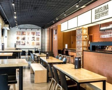 El moderno interior de la cervecería cuenta con mesas y sillas de madera y una acogedora zona de bar. Los amplios ventanales iluminan el cartel de Barcelona Beer Company, creando un ambiente ideal para disfrutar de la "Cata de cervezas y quesos artesanos" (Barcelona) con cervezas y quesos artesanales. - ForTwo - Planes para parejas