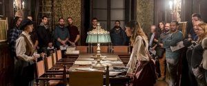 En una sala vintage, la gente se reúne alrededor de una gran mesa cubierta de papeles, cautivada por dos mujeres que desentrañan la Cena Enigma, que recuerda a la "Cena a lo Sherlock Holmes con la Cena Enigma de Sternalia" en Barcelona. - ForTwo - Planes para parejas