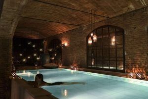 En una zona de piscina subterránea poco iluminada, con paredes de ladrillo y luces colgantes, que recuerda a la "Experiencia Baño termal con masaje en Aires de Barcelona", experimente momentos serenos de relajación. - ForTwo - Planes para parejas